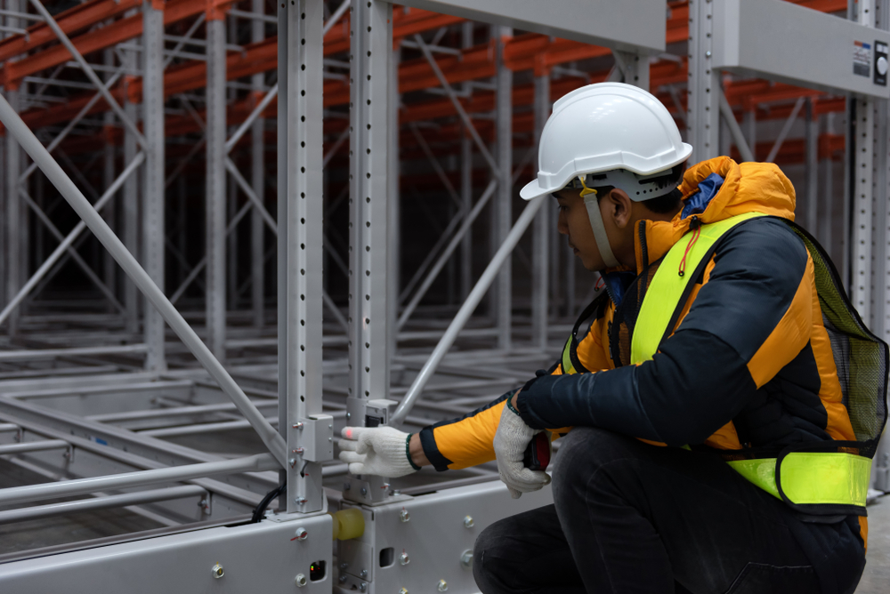 Worker in safety gear kneeling and inspecting a metal structure in an industrial warehouse, wearing a white hard hat, orange and black jacket, and yellow safety vest.